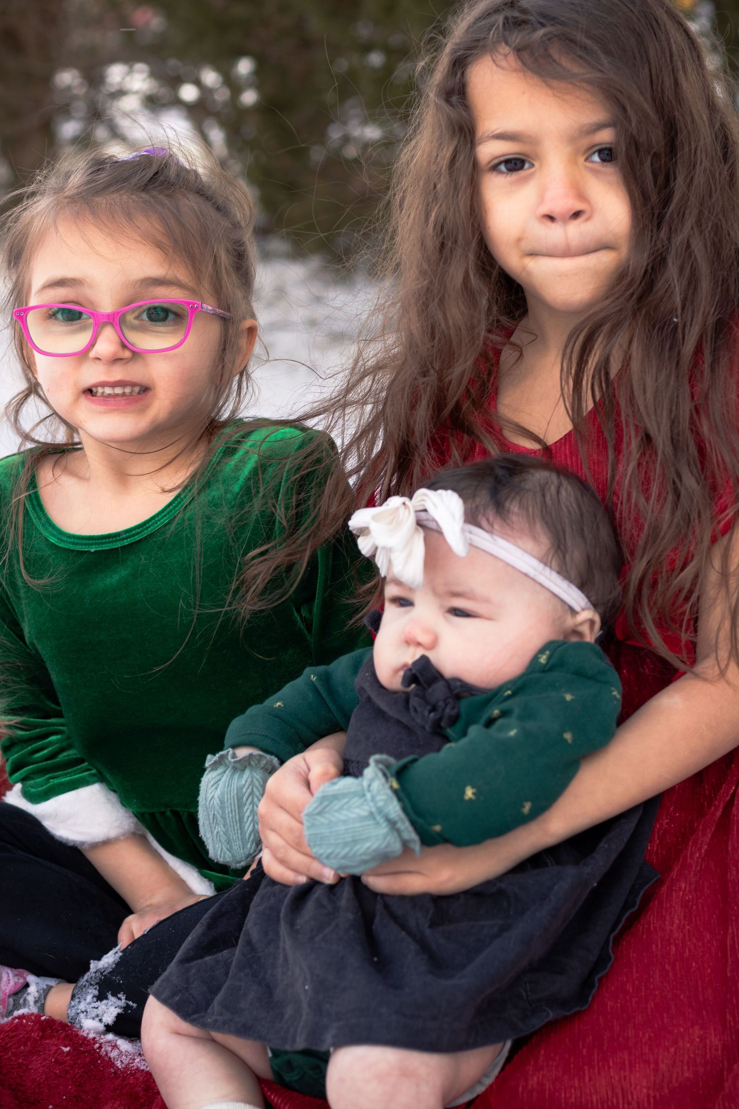 Two young sisters in green and red velvet dresses hold their baby sibling during a winter photo session.