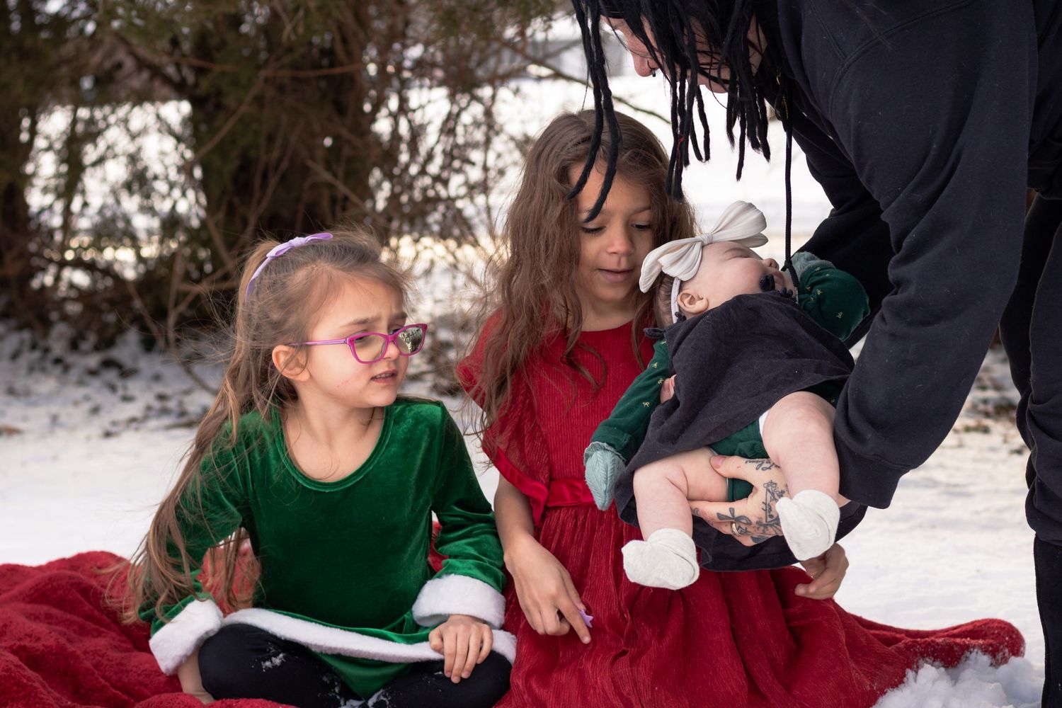 Two girls in holiday dresses sit in snow with grandma handing  baby to sister, surrounded by winter scenery.