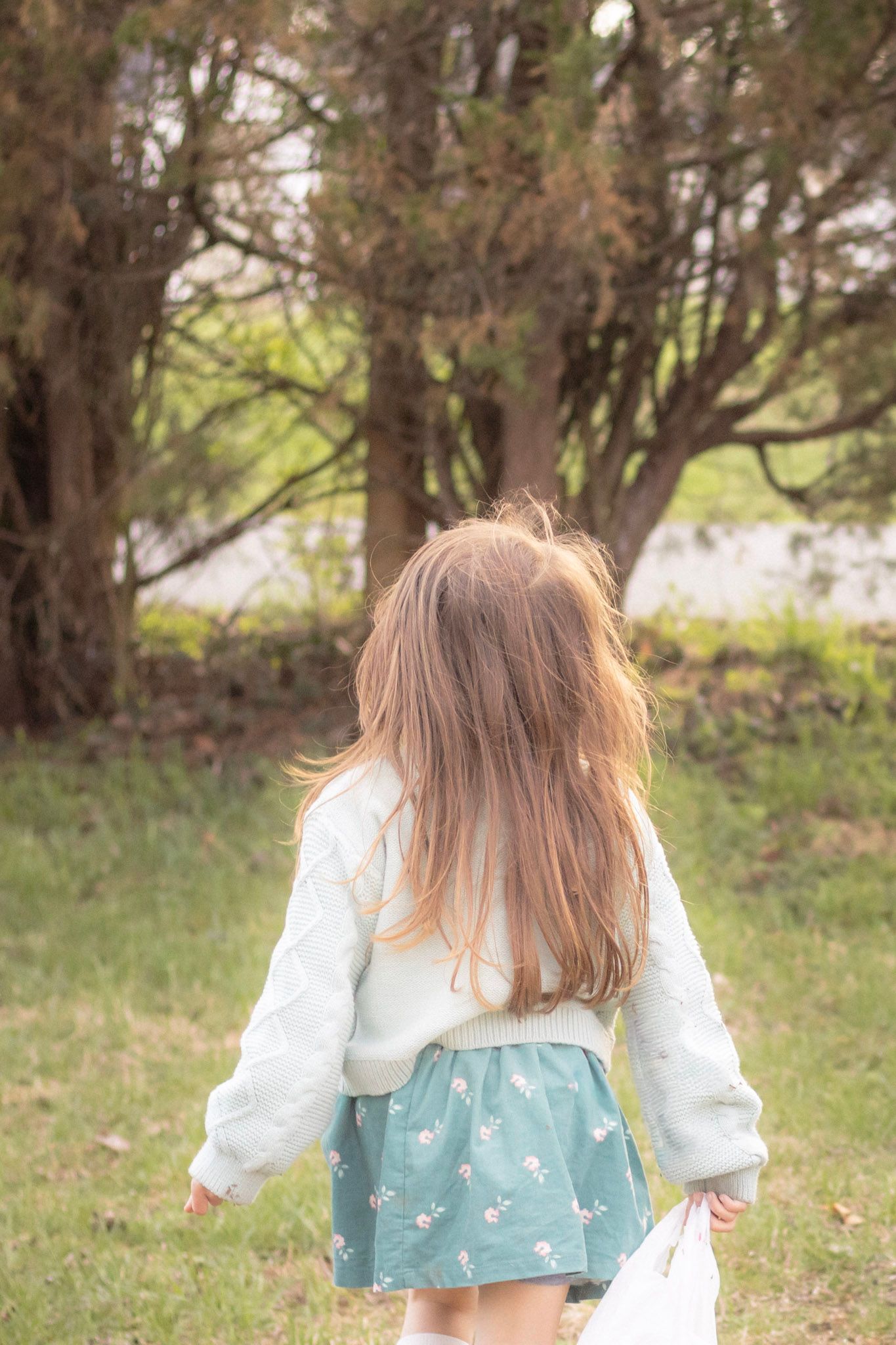 Young girl with long hair in floral skirt and white sweater walking toward trees in a green park.