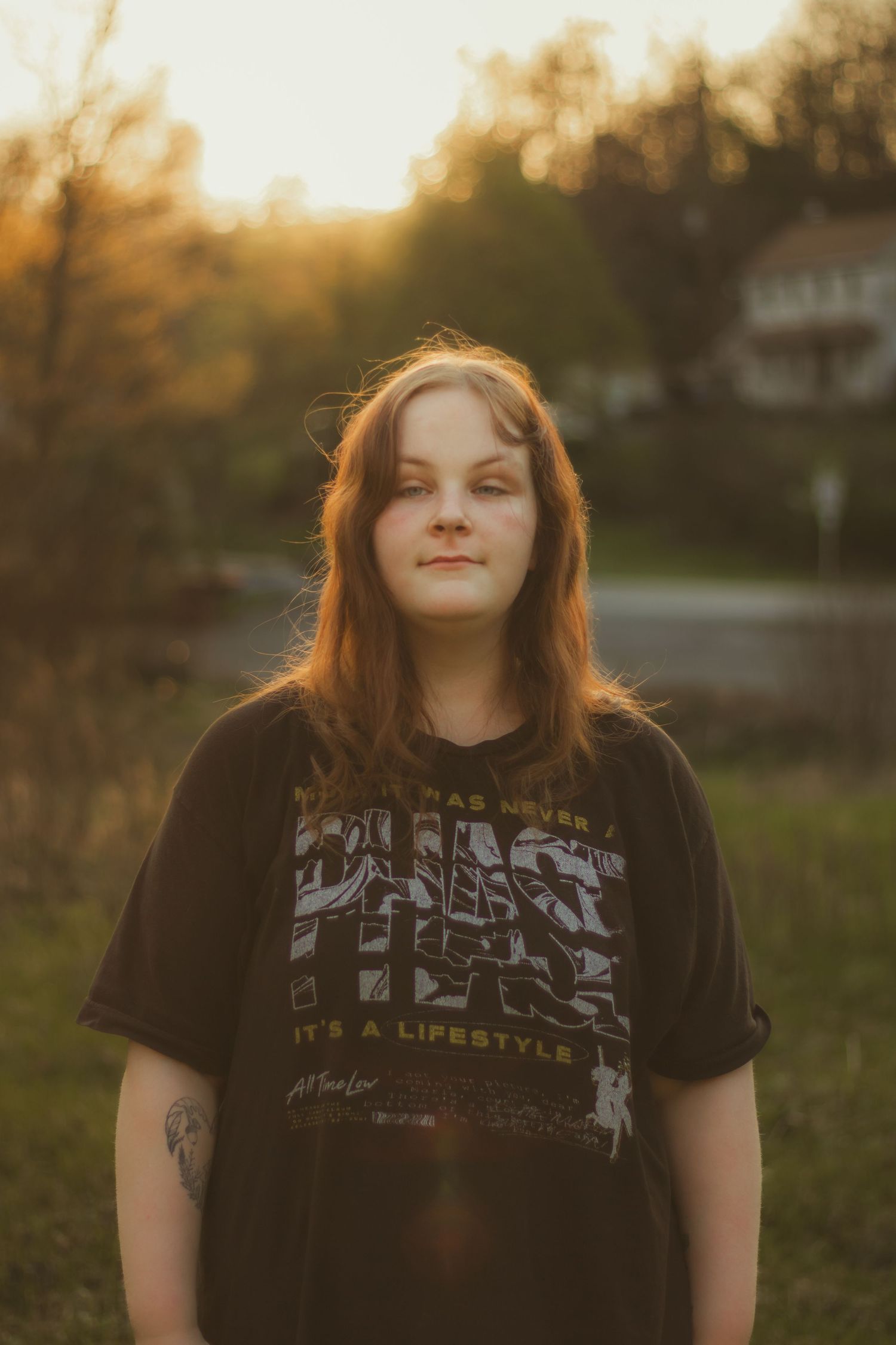 Young woman with brown hair stands confidently outdoors at sunset wearing a black Phish graphic tee with tattoos visible.