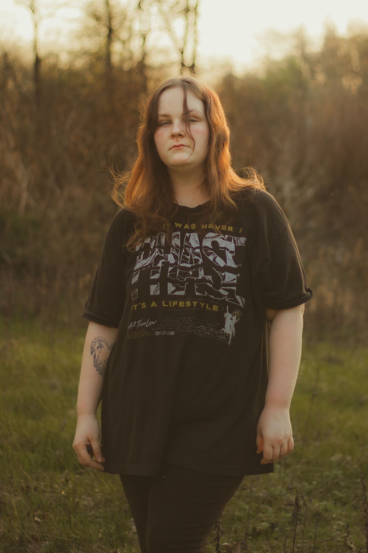 Young woman with auburn hair stands in a grassy field at sunset wearing an oversized black Phish graphic tee.
