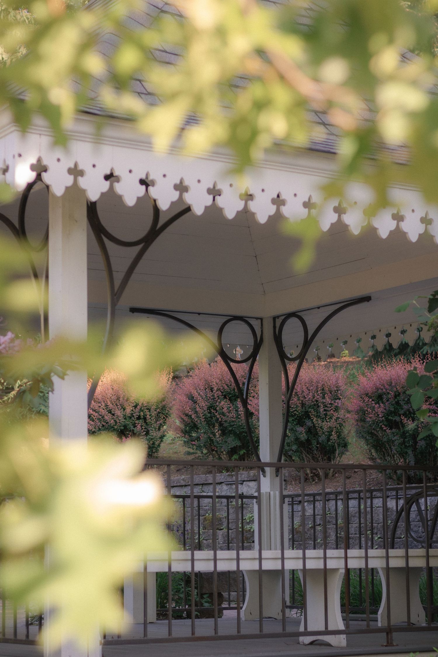 White ornate gazebo with decorative trim and iron railings, surrounded by lush green trees and purple flowering shrubs.