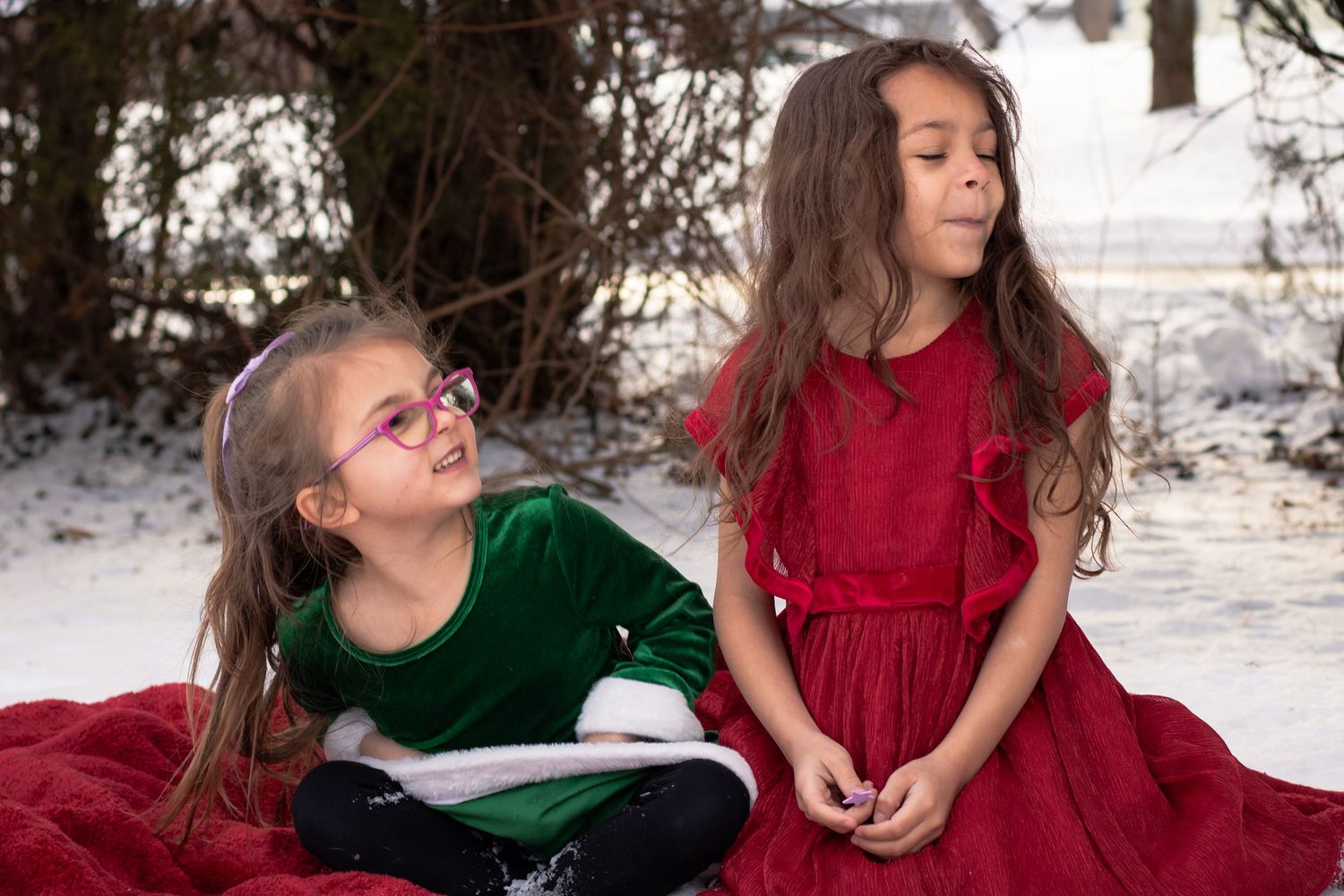 Two young girls in green and red holiday dresses sit together on a red blanket in a snowy landscape.