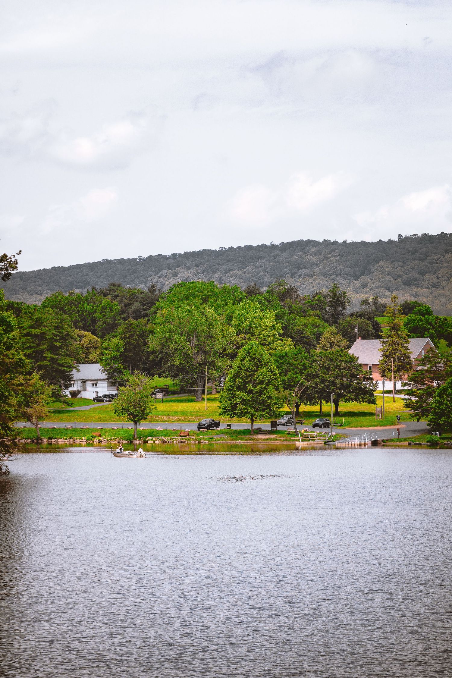 Scenic lakeside view with green trees, white farmhouse, and rolling hills under cloudy sky.