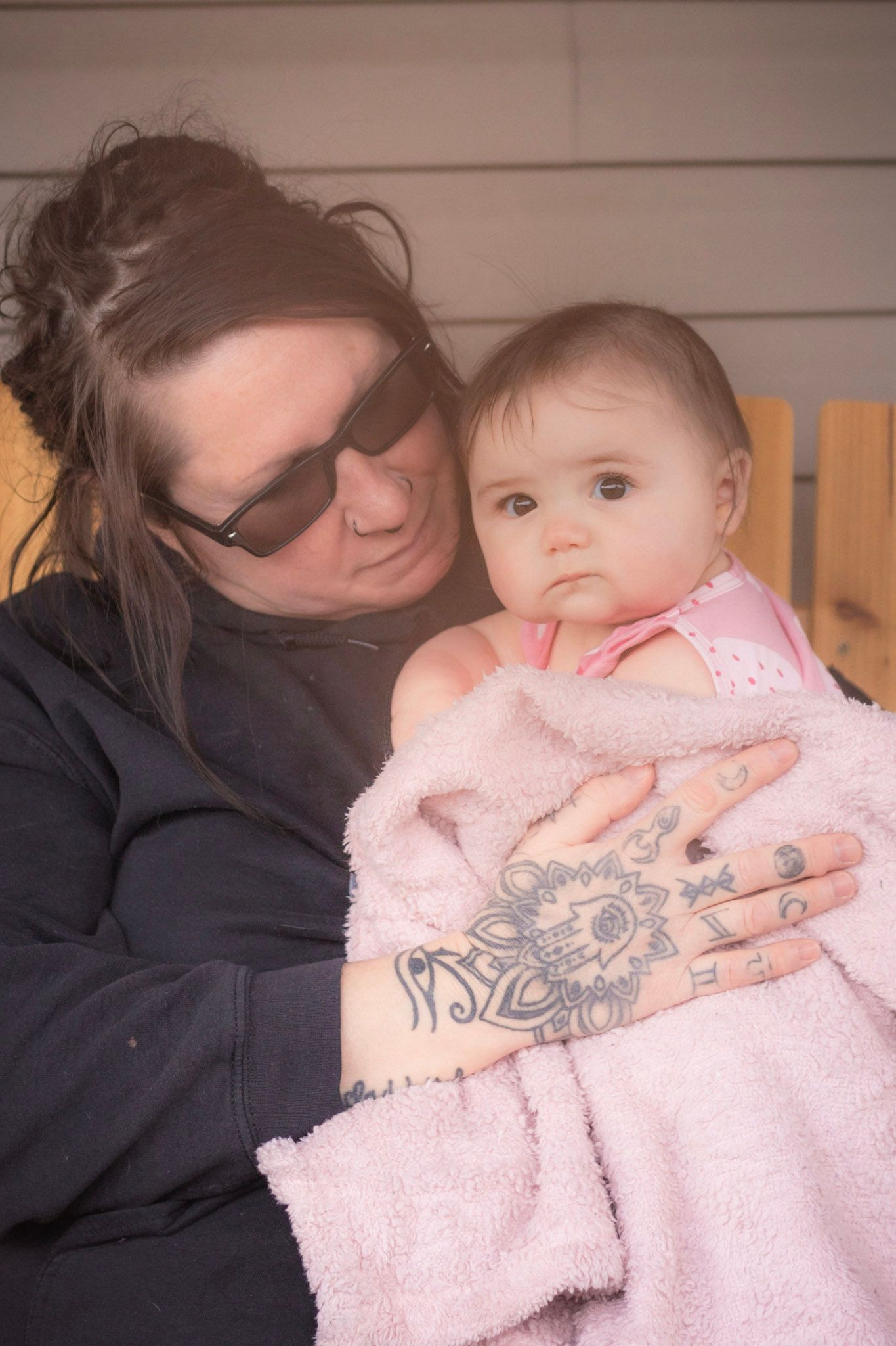 Tattooed woman in sunglasses tenderly holding a baby wrapped in a soft pink blanket outdoors.
