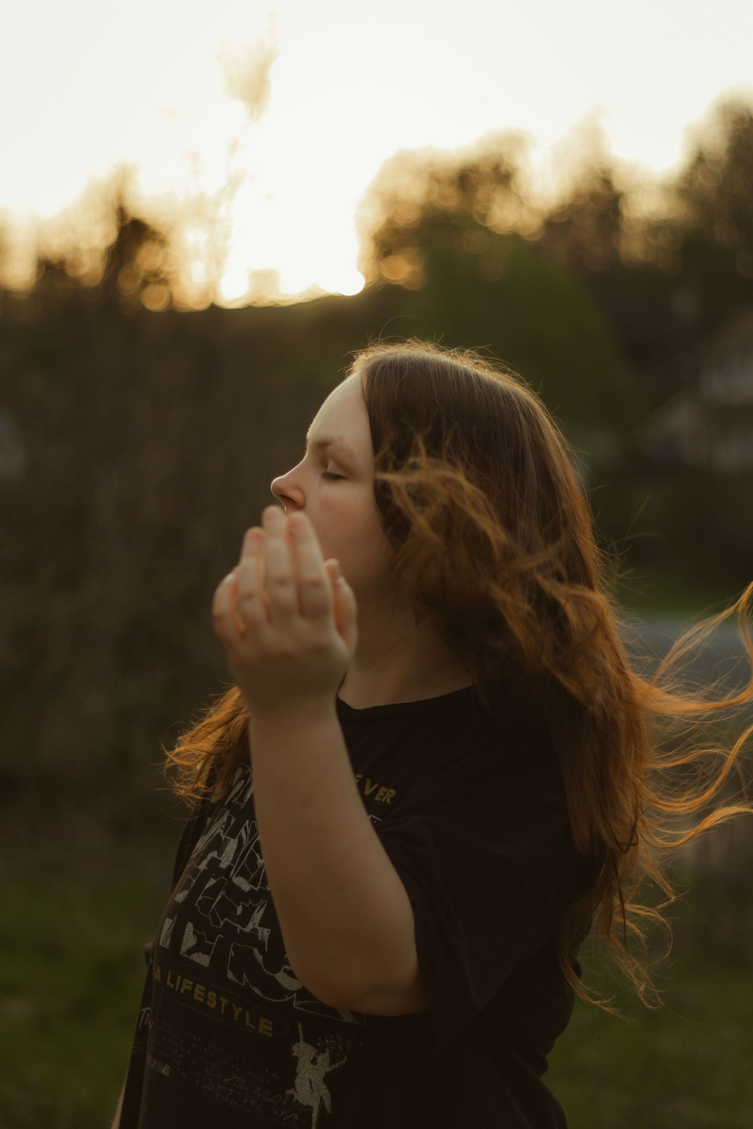 Young woman with windswept auburn hair raises her hand toward camera during a golden hour outdoor portrait session.