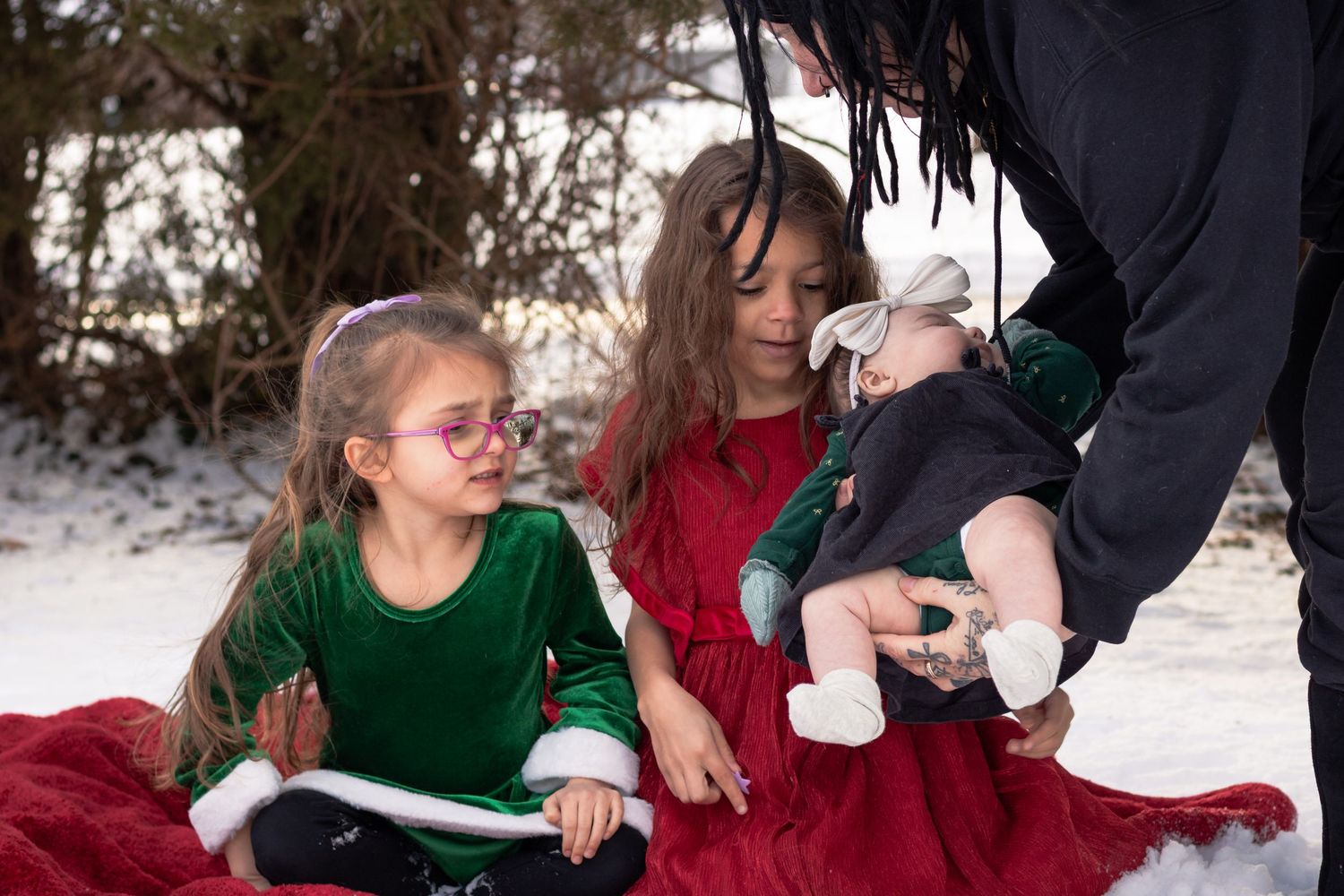 Two young girls in holiday dresses sit in snow with an adult holding a baby dressed in green.