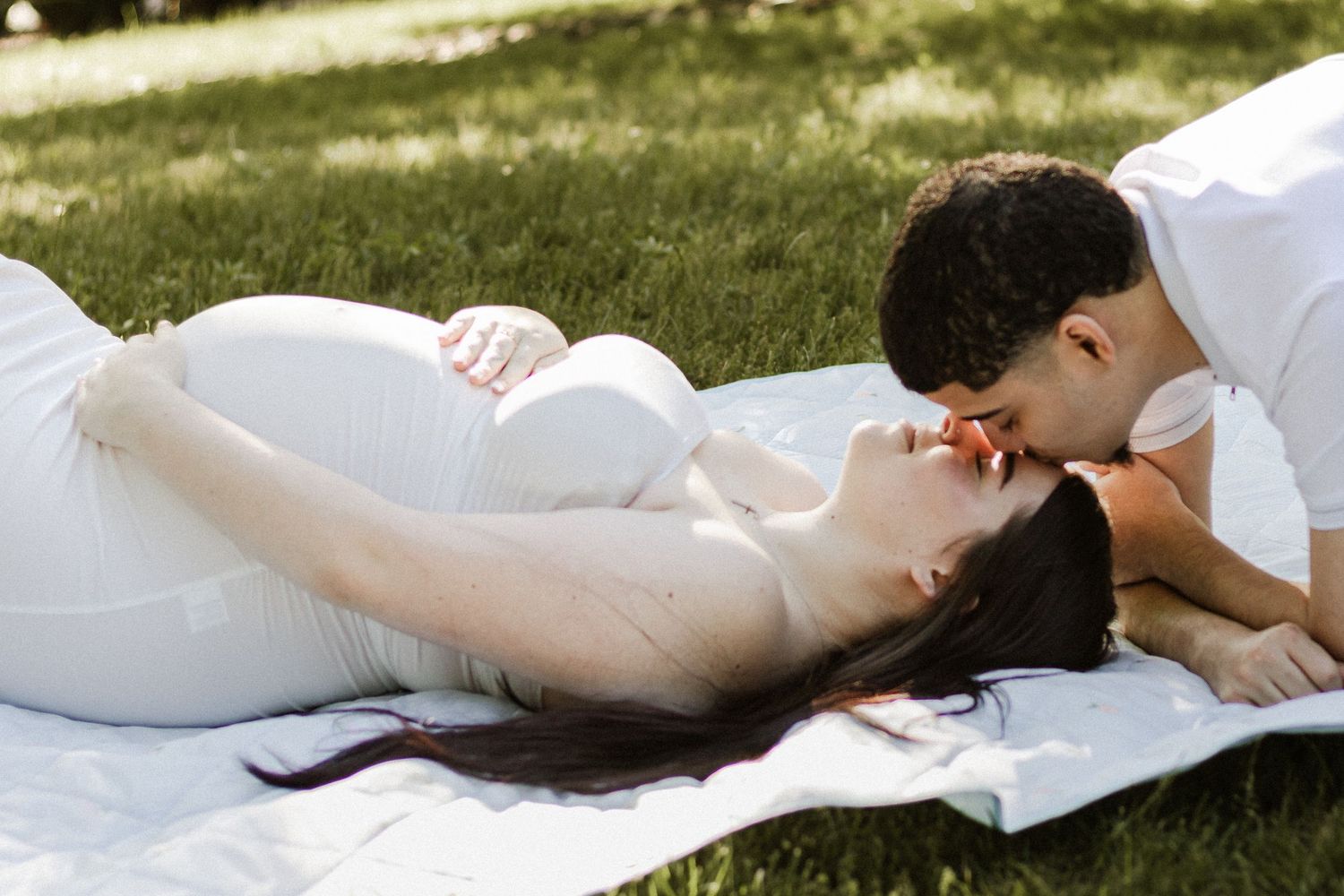 Pregnant woman in white dress lying on blanket in park, sharing a tender kiss with her partner.
