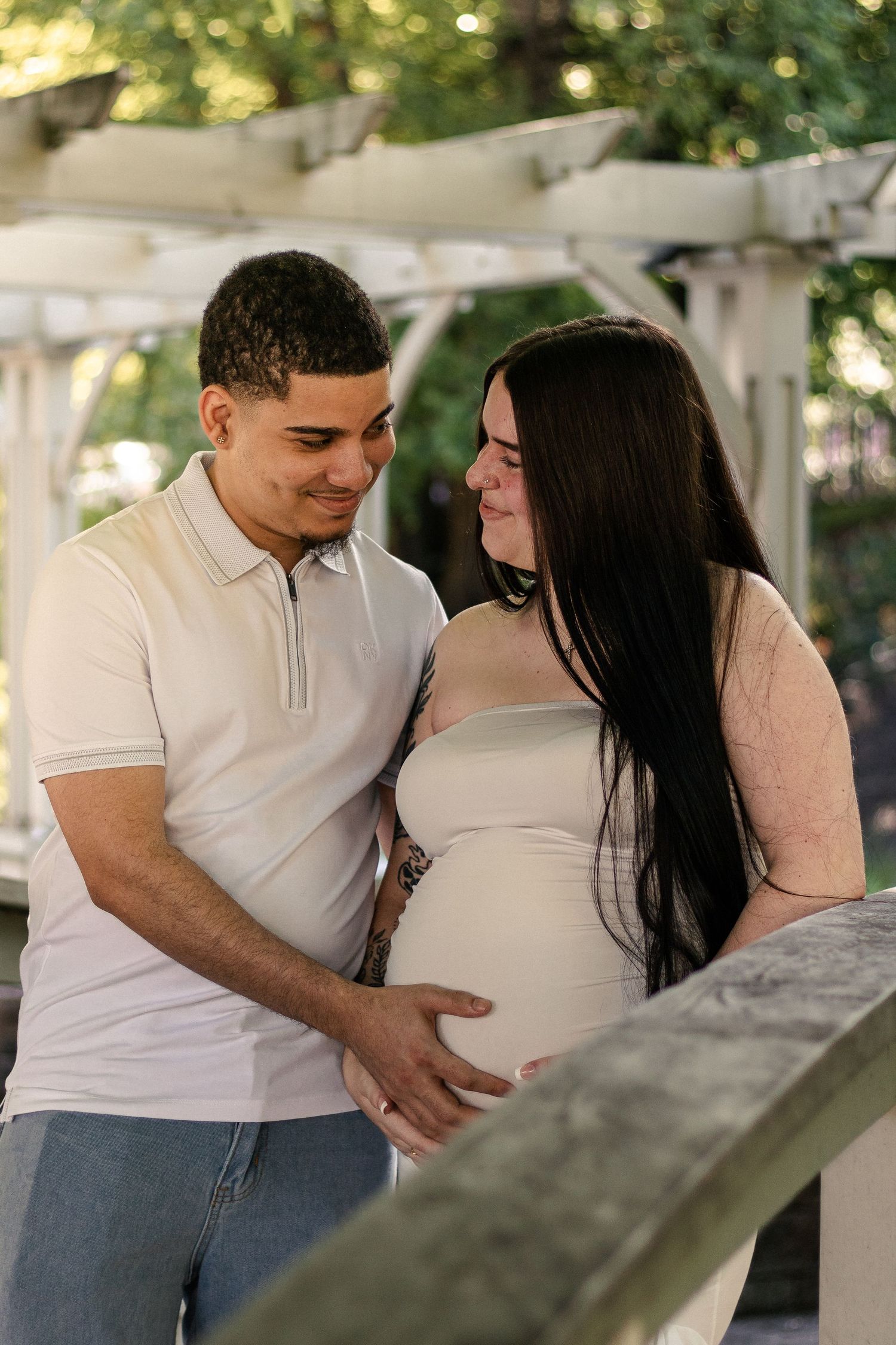 A couple tenderly cradles a pregnant belly outdoors under a white pergola surrounded by lush greenery.