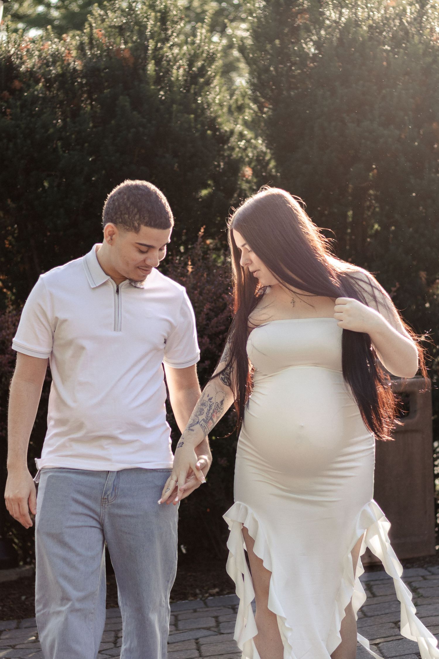 A couple walks outdoors, the pregnant woman in a white dress holding hands with her partner in a white polo shirt.
