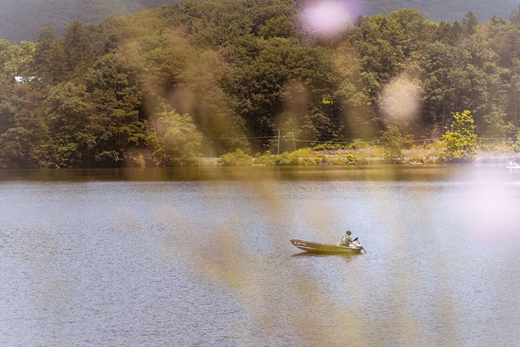 A lone fisherman rowing a small boat across a calm lake surrounded by lush green forest.