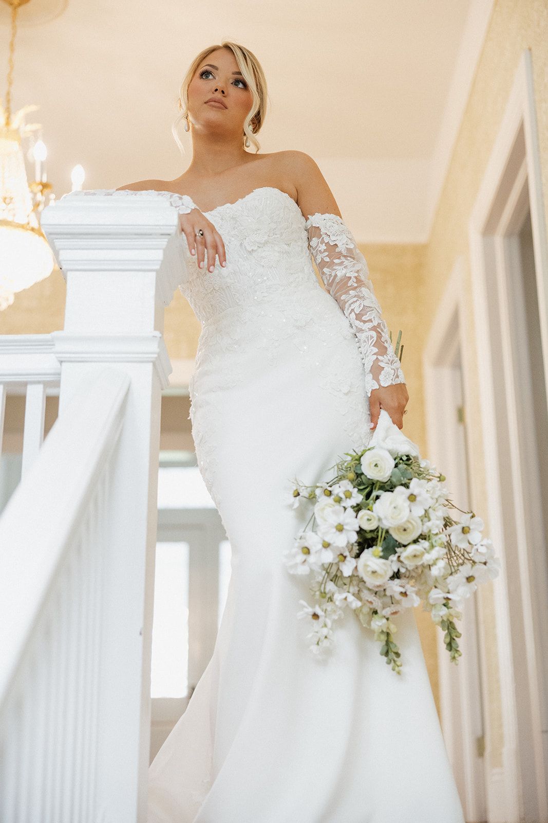 Elegant bride in white lace gown with long sleeves poses by a classic white pillar holding a white floral bouquet.