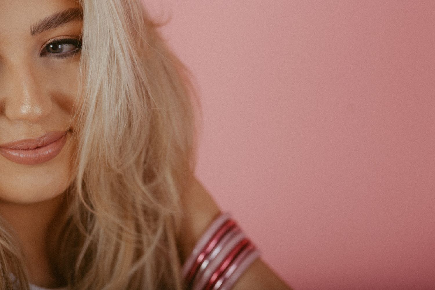 Close-up cropped portrait against pink background showing blonde wavy hair and partial smile.