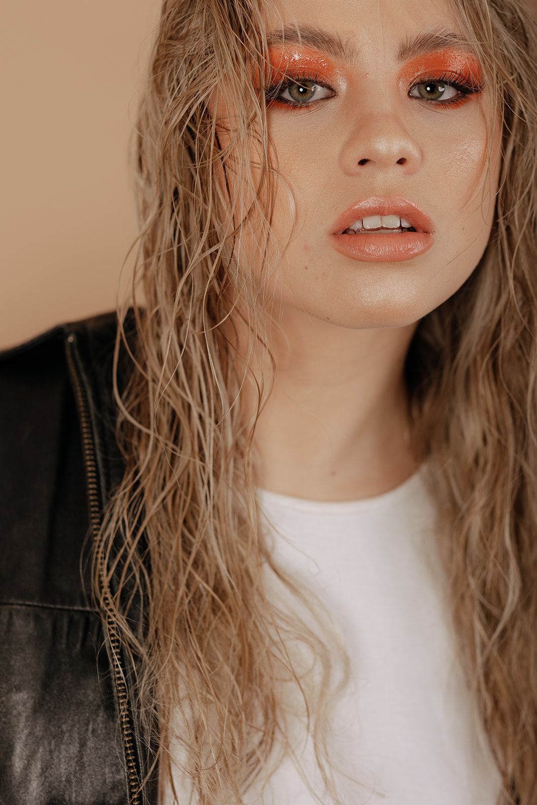 Stylish beauty look featuring vibrant orange eyeshadow, wavy blonde hair, and a leather jacket with white top.