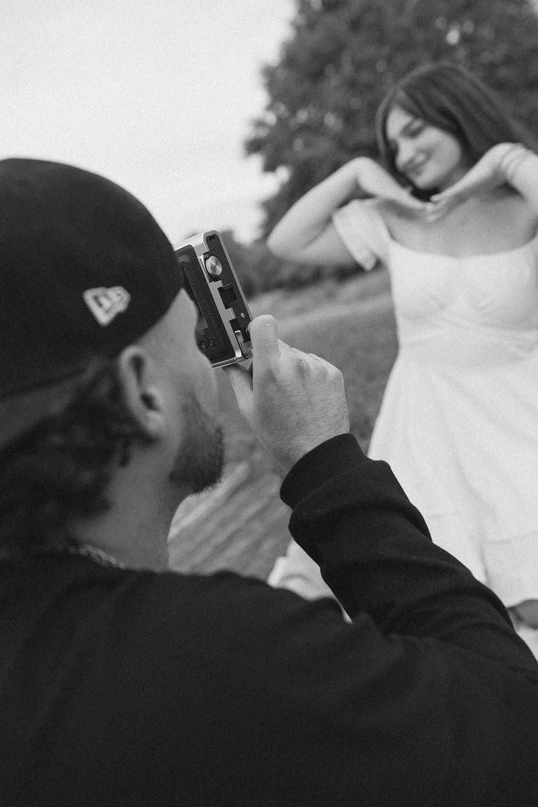 A photographer with a vintage camera captures images of a woman in a white dress against a natural backdrop.