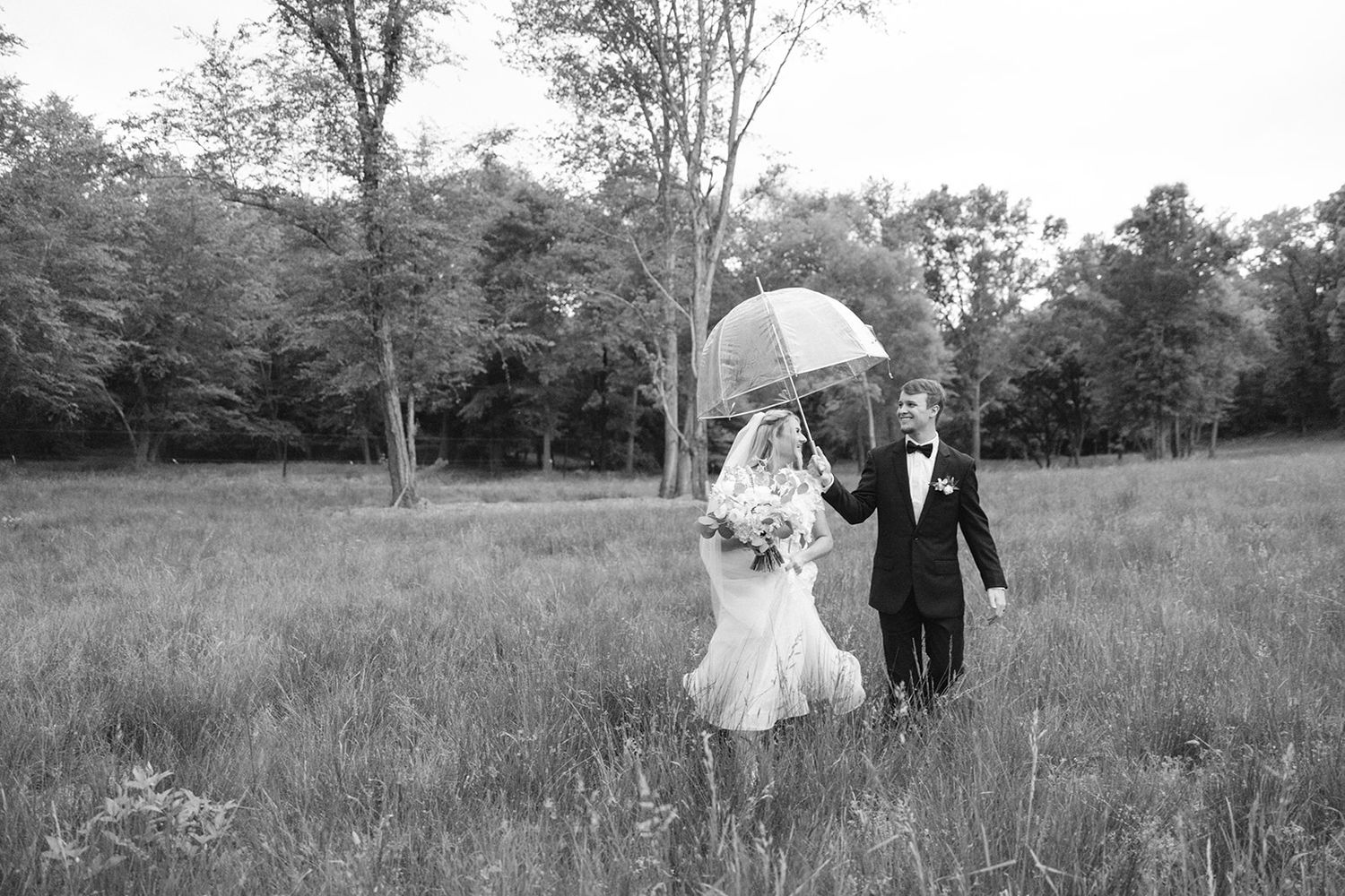 Couple walking through a meadow with white umbrella during wedding photo session in black and white.