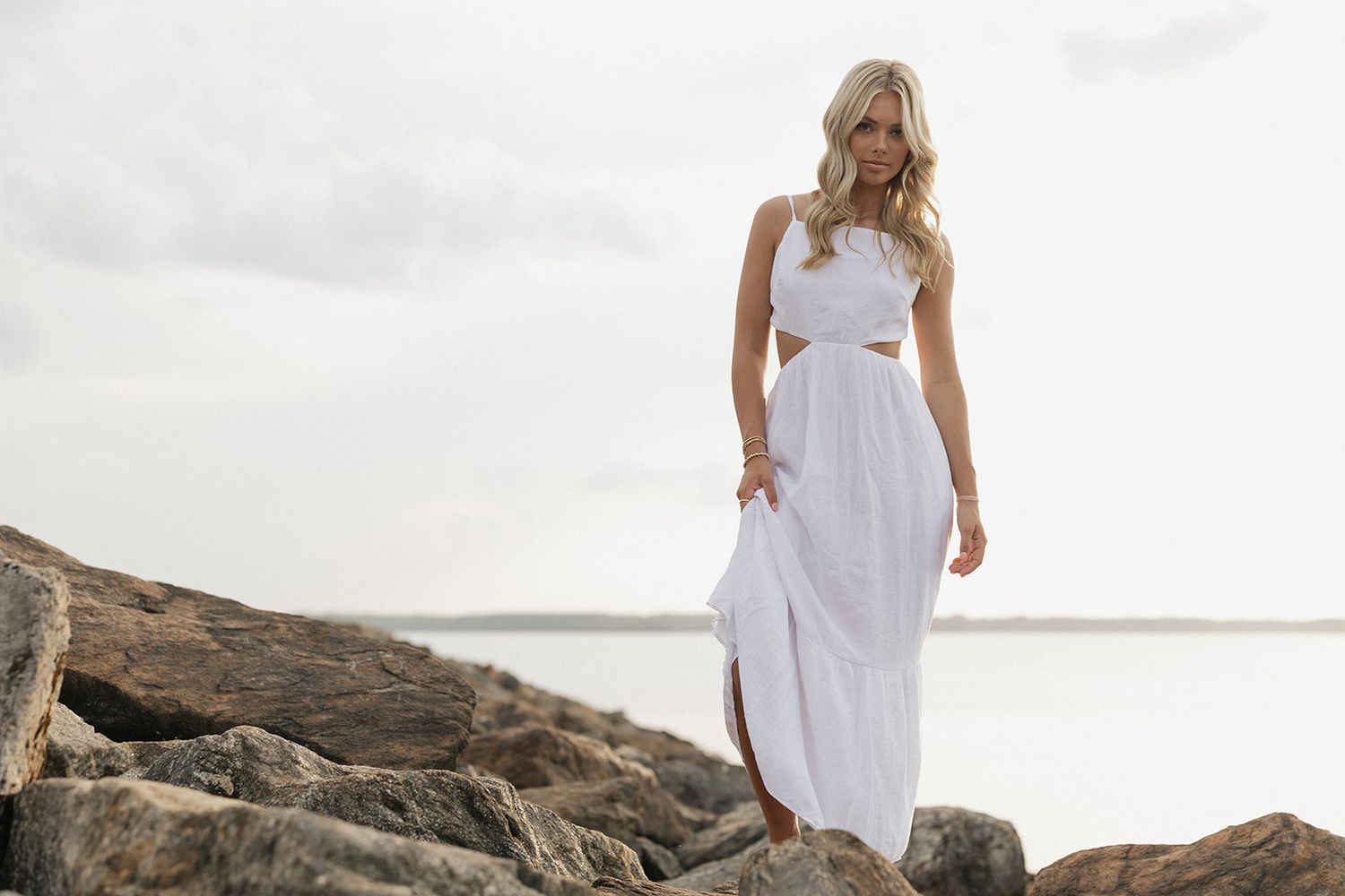 Person in flowing white dress walking gracefully along scenic rocky coastline at sunset.