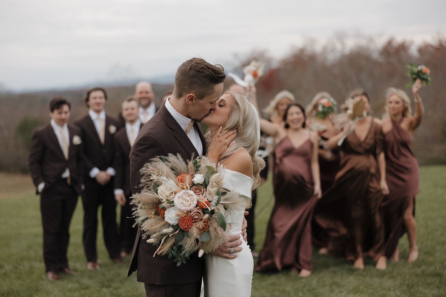 Wedding party in dark formal attire posing together on grassy field during outdoor celebration.