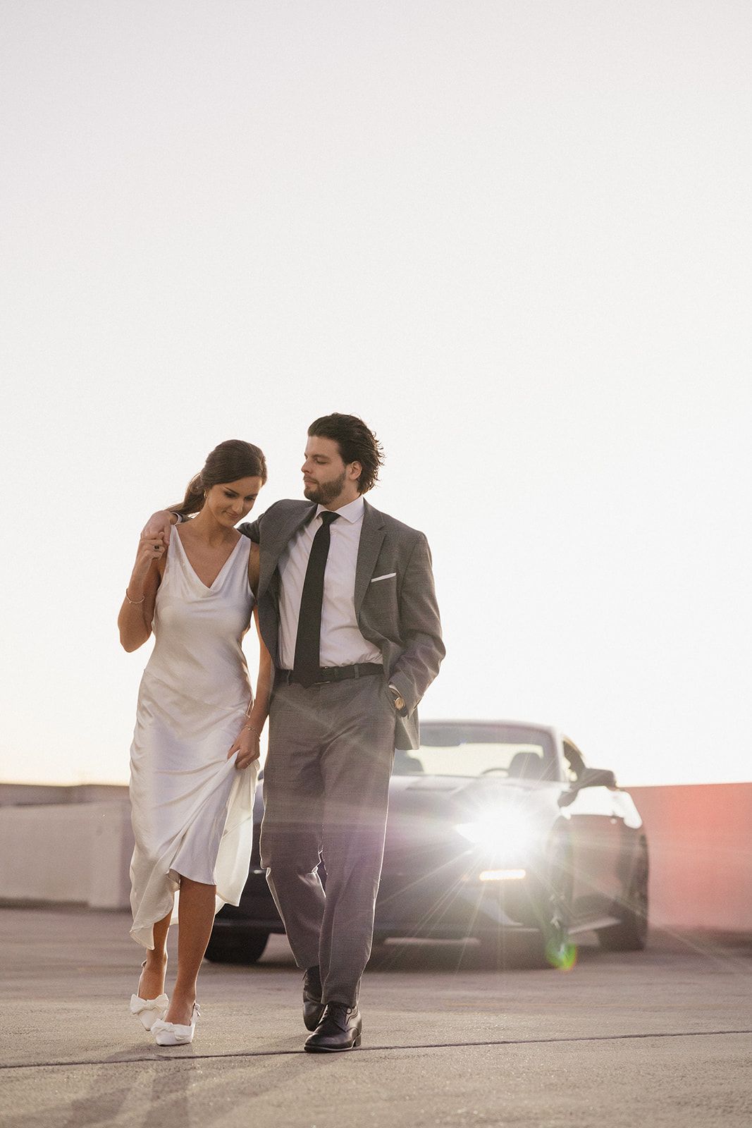 Couple walks hand in hand past parked car during sunset photoshoot.