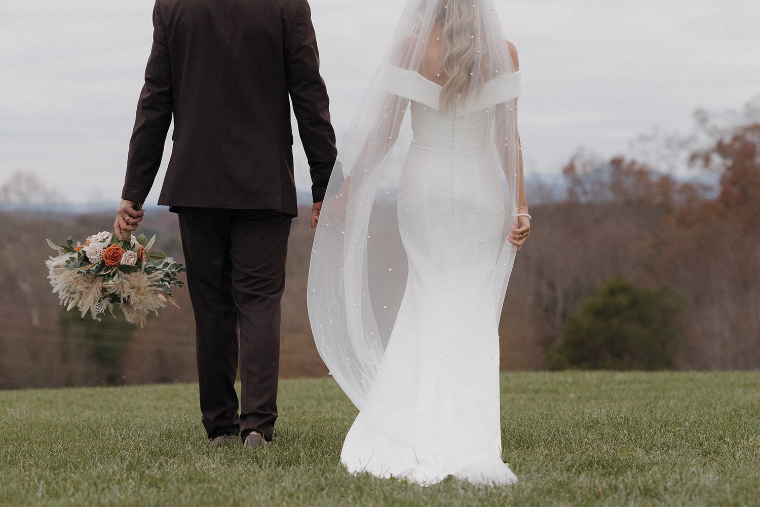 Wedding couple walking hand in hand through an open green field in formal attire.