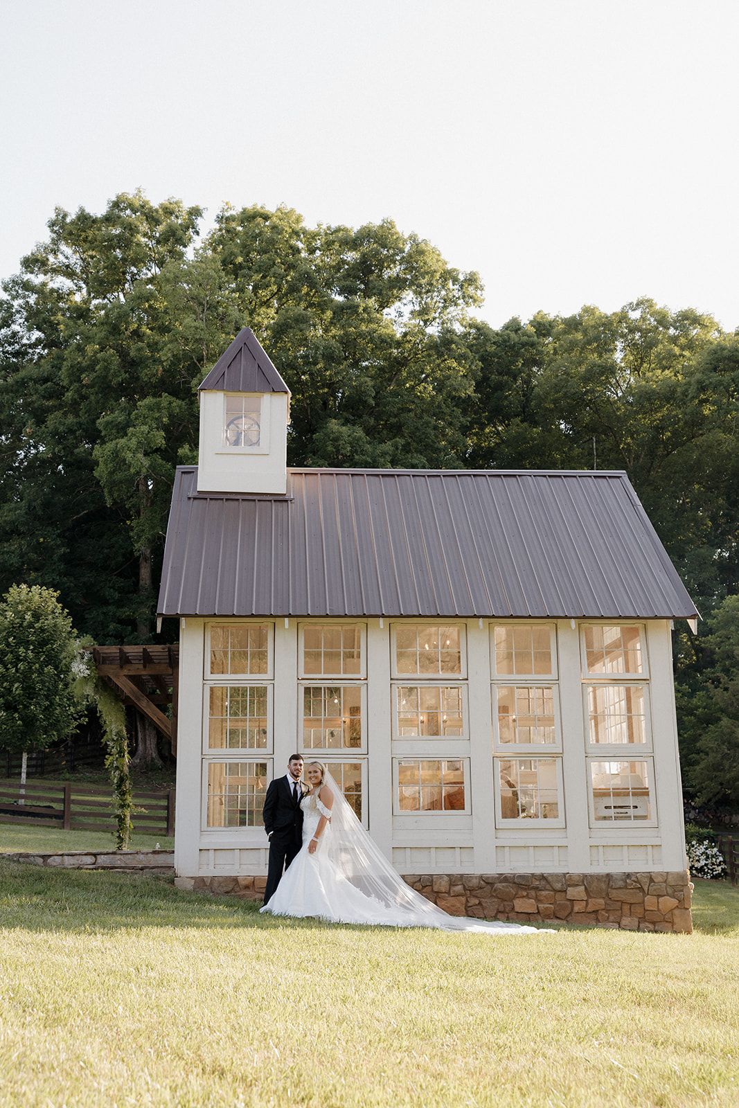 Wedding couple poses in front of charming white chapel with glass windows at sunset.