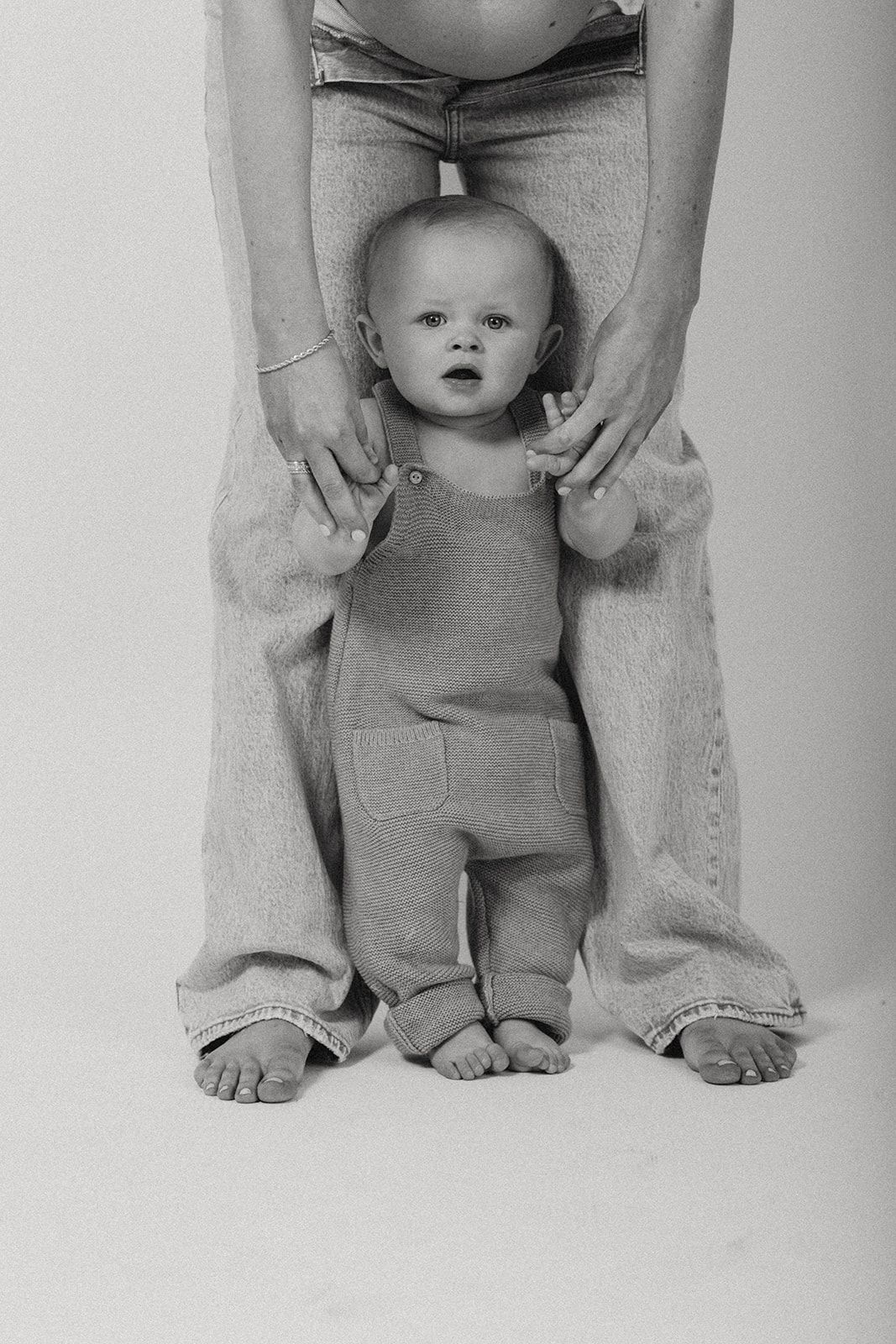 Black and white photo of a baby in overalls standing between adult legs in denim jeans against white background.