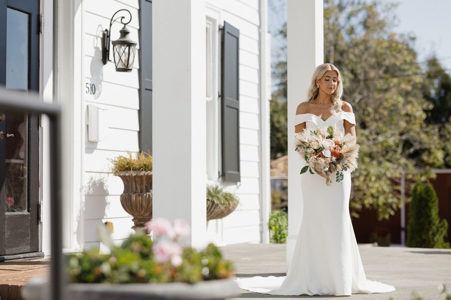 Beautiful bride in fitted white lace gown holds floral bouquet while standing outside elegant white mansion with lanterns.