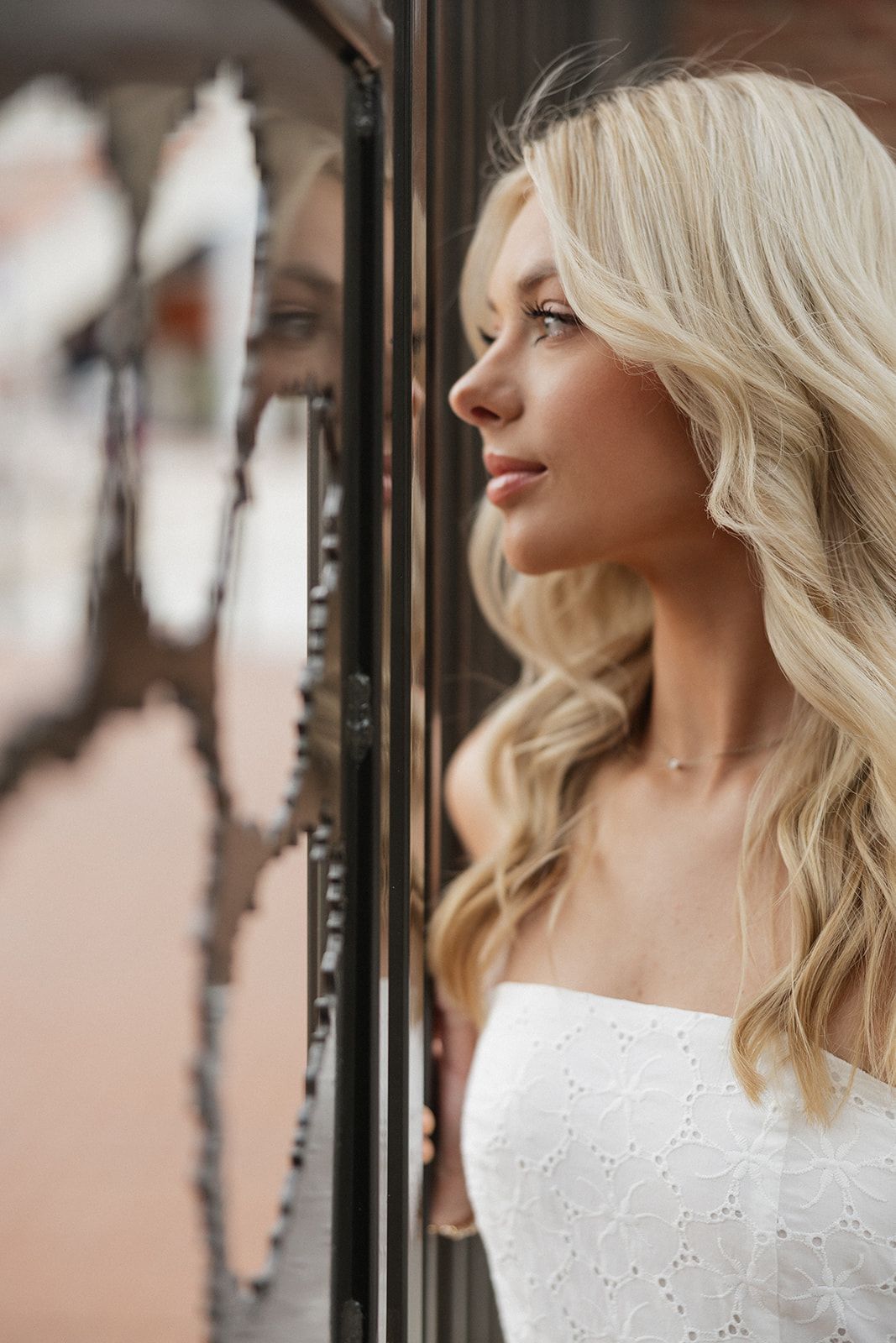 Side profile of blonde woman in white eyelet dress gazing through window with soft lighting and blurred background.