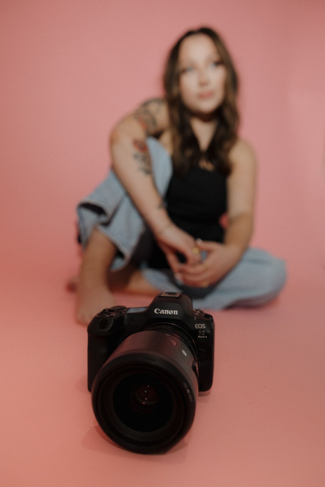 Canon camera with large lens in foreground while a seated figure in black and denim appears blurred in pink background.