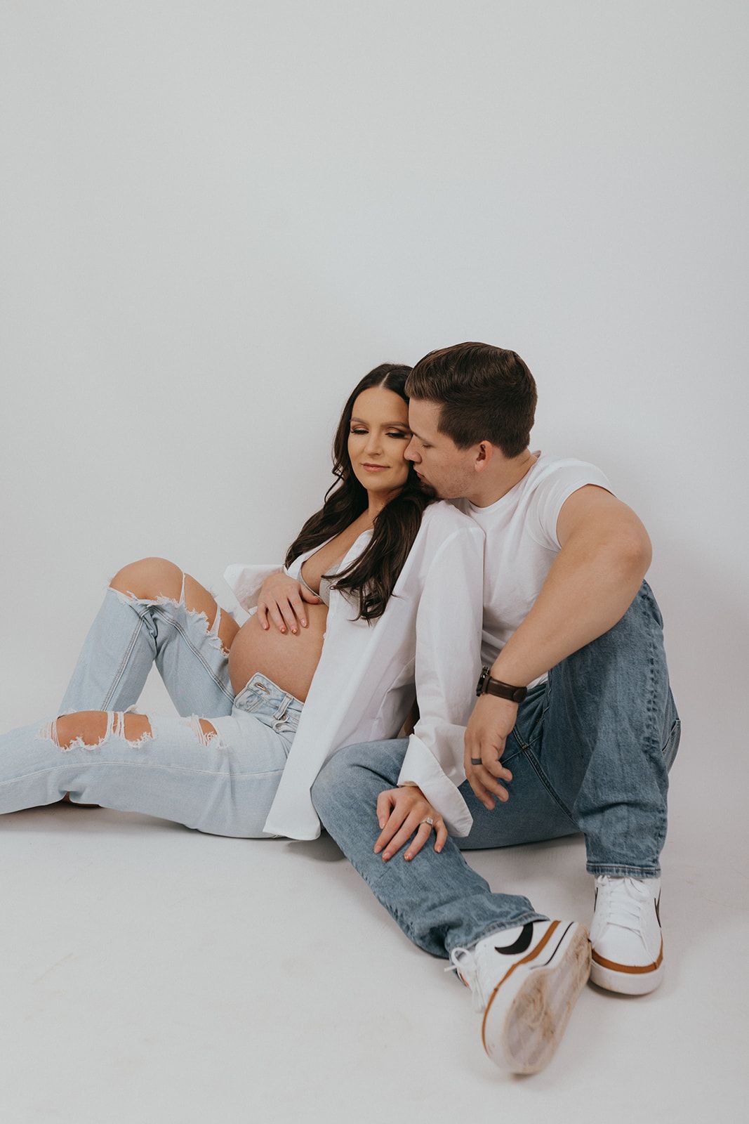 Two people in casual denim and white tops sitting close together on white studio floor.