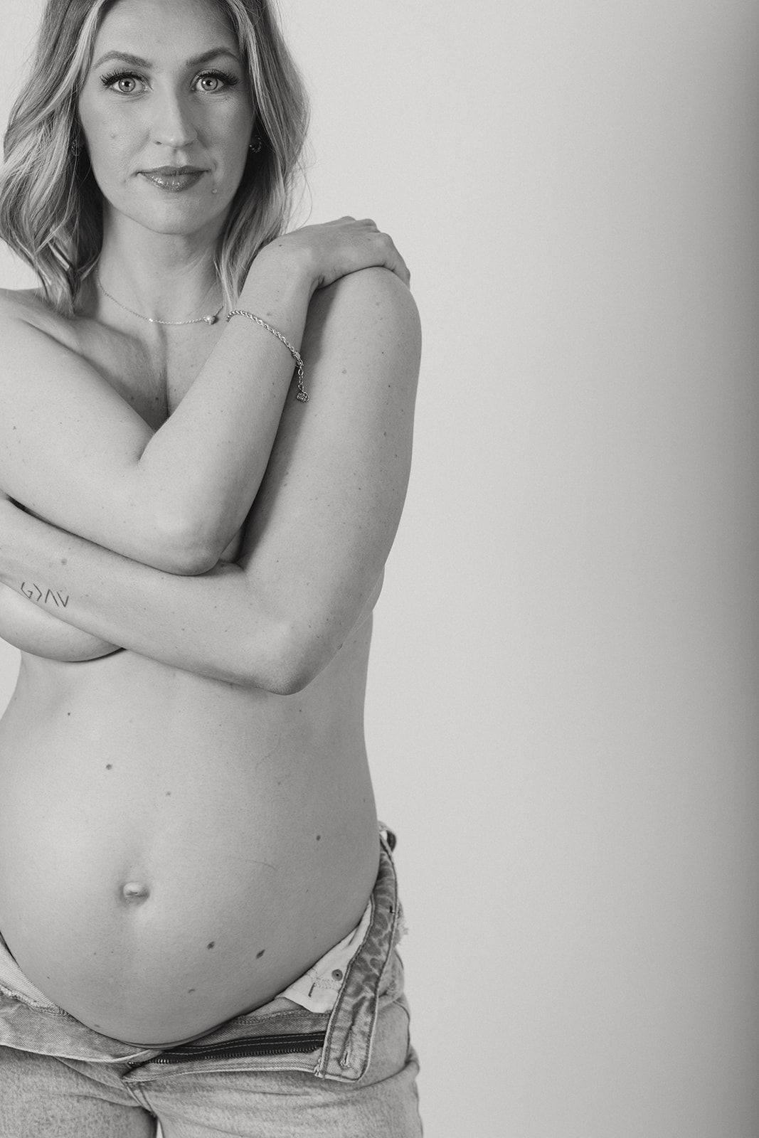 Black and white artistic maternity photo showing a bare belly and crossed arms in an intimate natural pose with jeans.