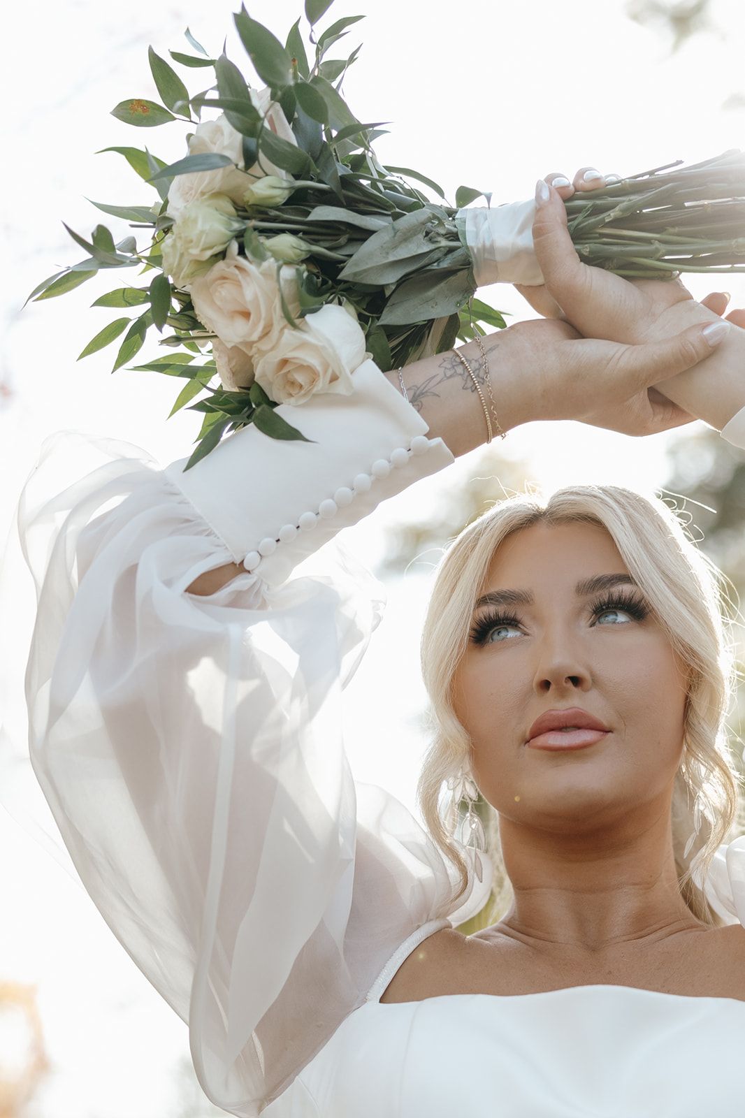 Bride holding romantic white rose bouquet with eucalyptus leaves against sunlit background.