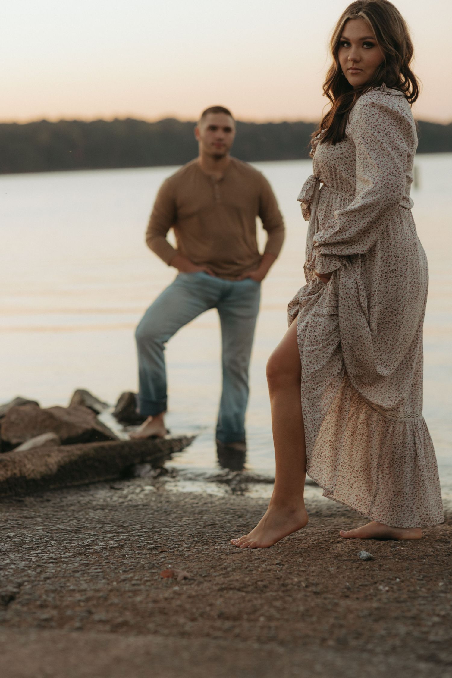 Stylish couple poses by lake at sunset, woman in lace dress with man standing on rocks.
