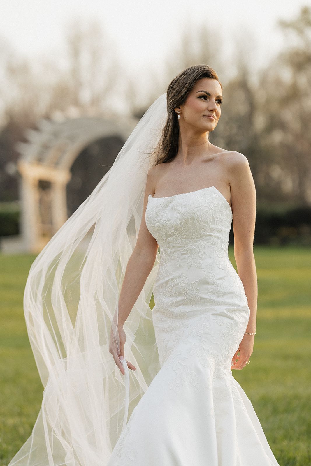 Bride in flowing white wedding dress and veil stands gracefully in outdoor setting during golden hour.