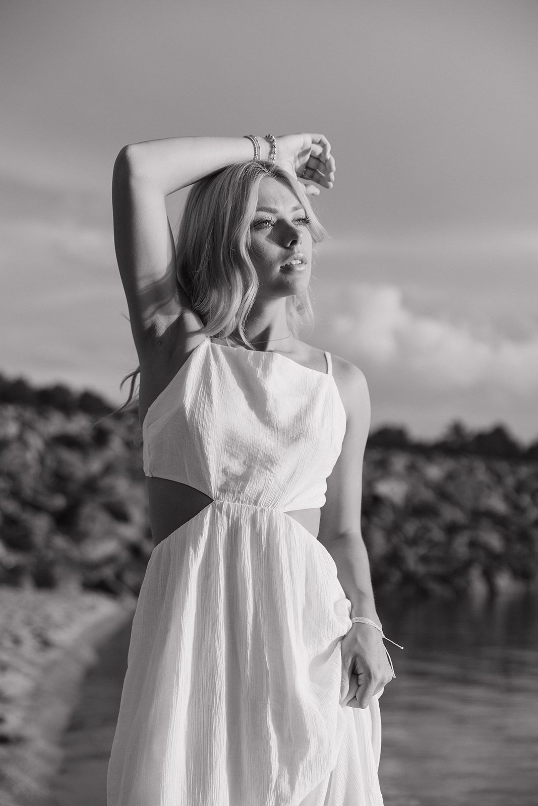 Black and white portrait of a person in a flowing white dress posing elegantly by a waterfront.
