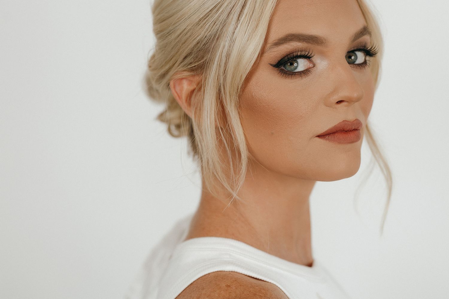Glamorous bridal beauty look with blonde updo hairstyle, smokey eye makeup and white dress against neutral background.