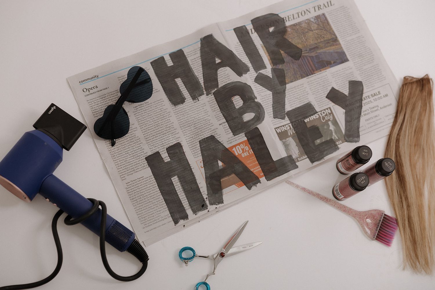 Hairdressing supplies including scissors, hair dryer, and text 'HAIR BY' arranged on gray newspaper background.