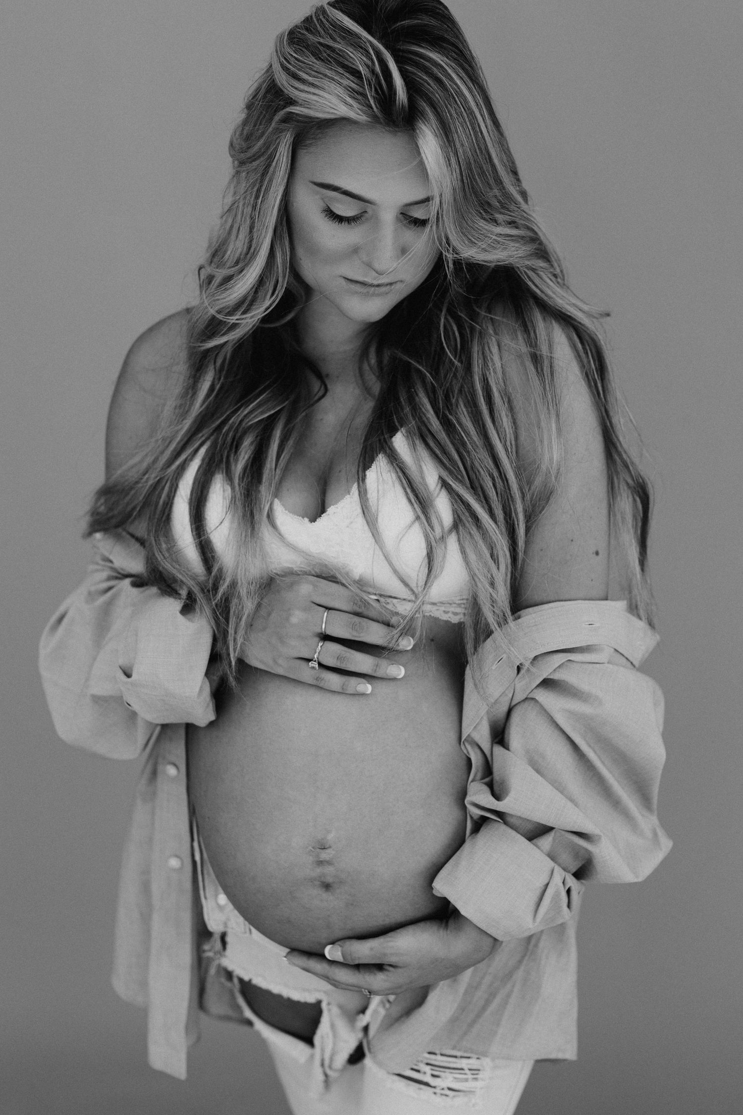 Black and white maternity photo showing a pregnant woman in an open button-down shirt gently cradling her bare belly.