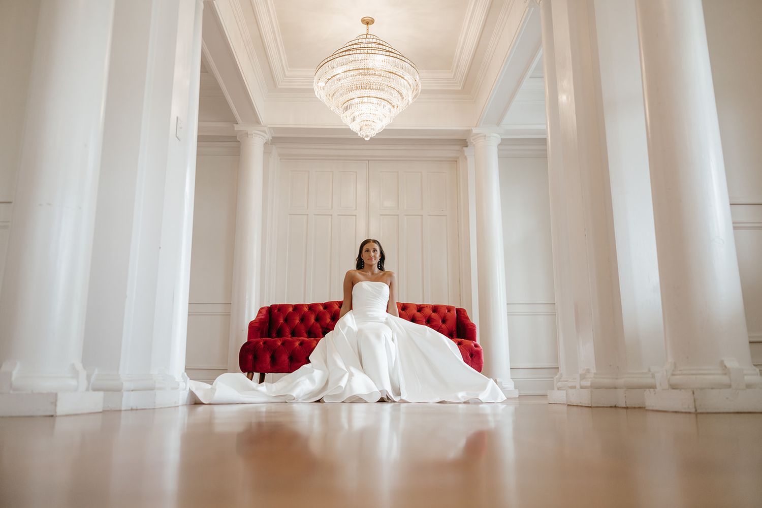 Minimalist white ballroom interior with crystal chandelier and red accent furniture.