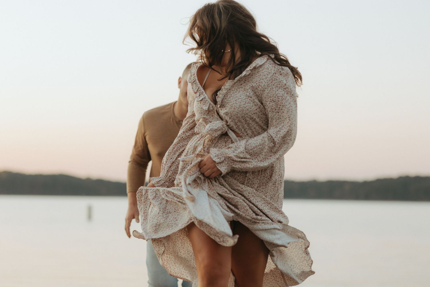 Intimate moment captured between couple by lakeside with flowing dress in evening light.
