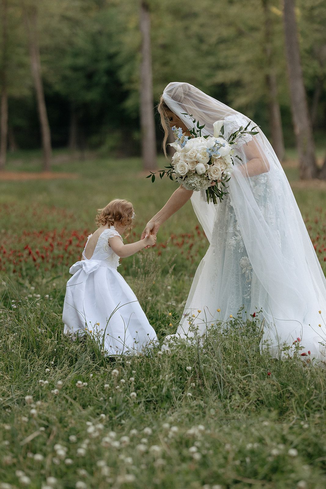 Bride in flowing white gown bends down to interact with flower girl in matching dress among wildflowers.