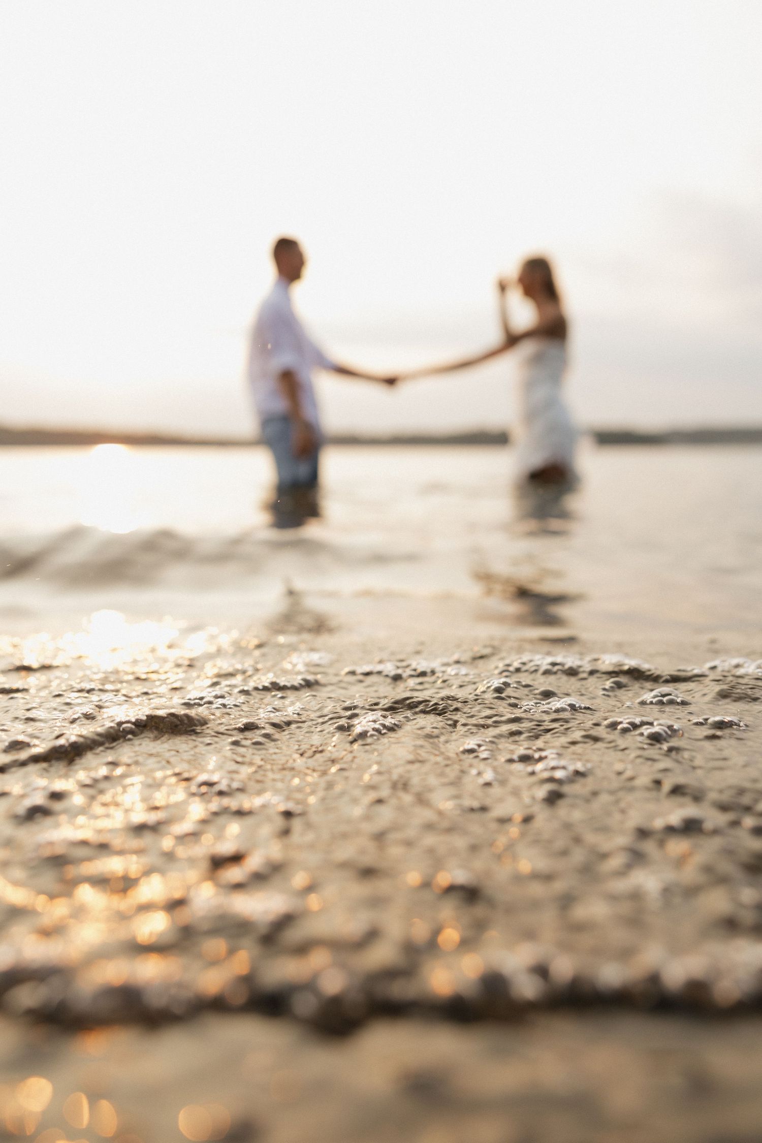 Soft focus shot of couple holding hands while standing in ocean water at sunset.