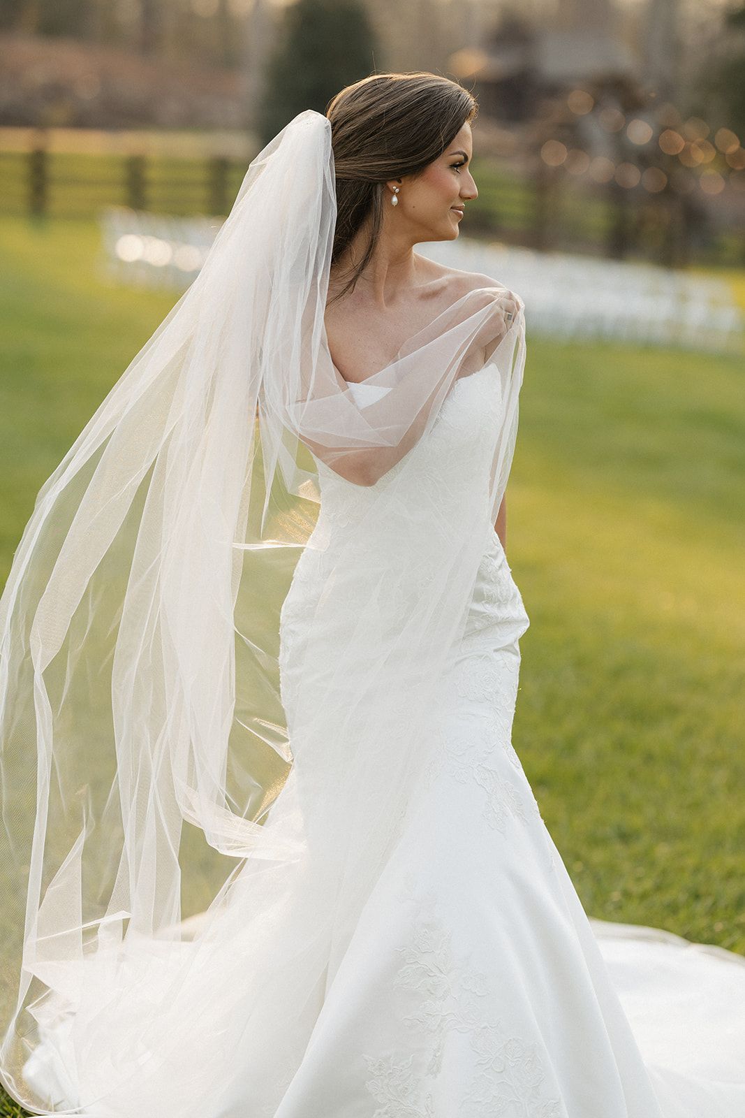Elegant bride in white mermaid wedding gown with flowing veil stands gracefully in sunlit garden setting.