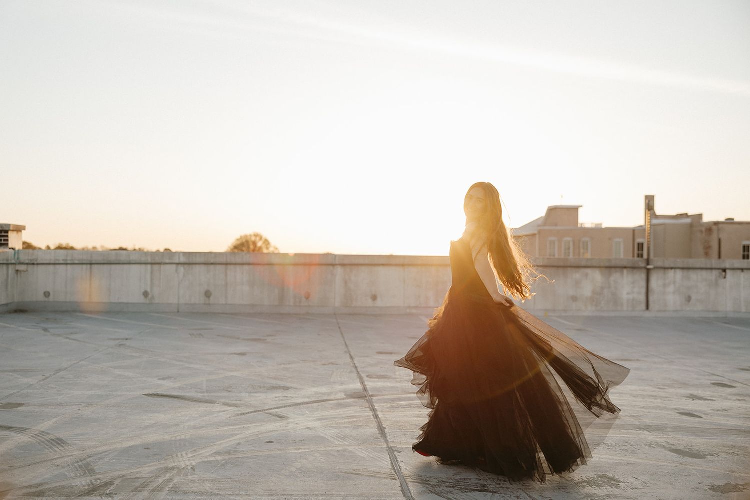 A figure in a flowing black dress twirls on a rooftop during golden hour with city buildings in the background.