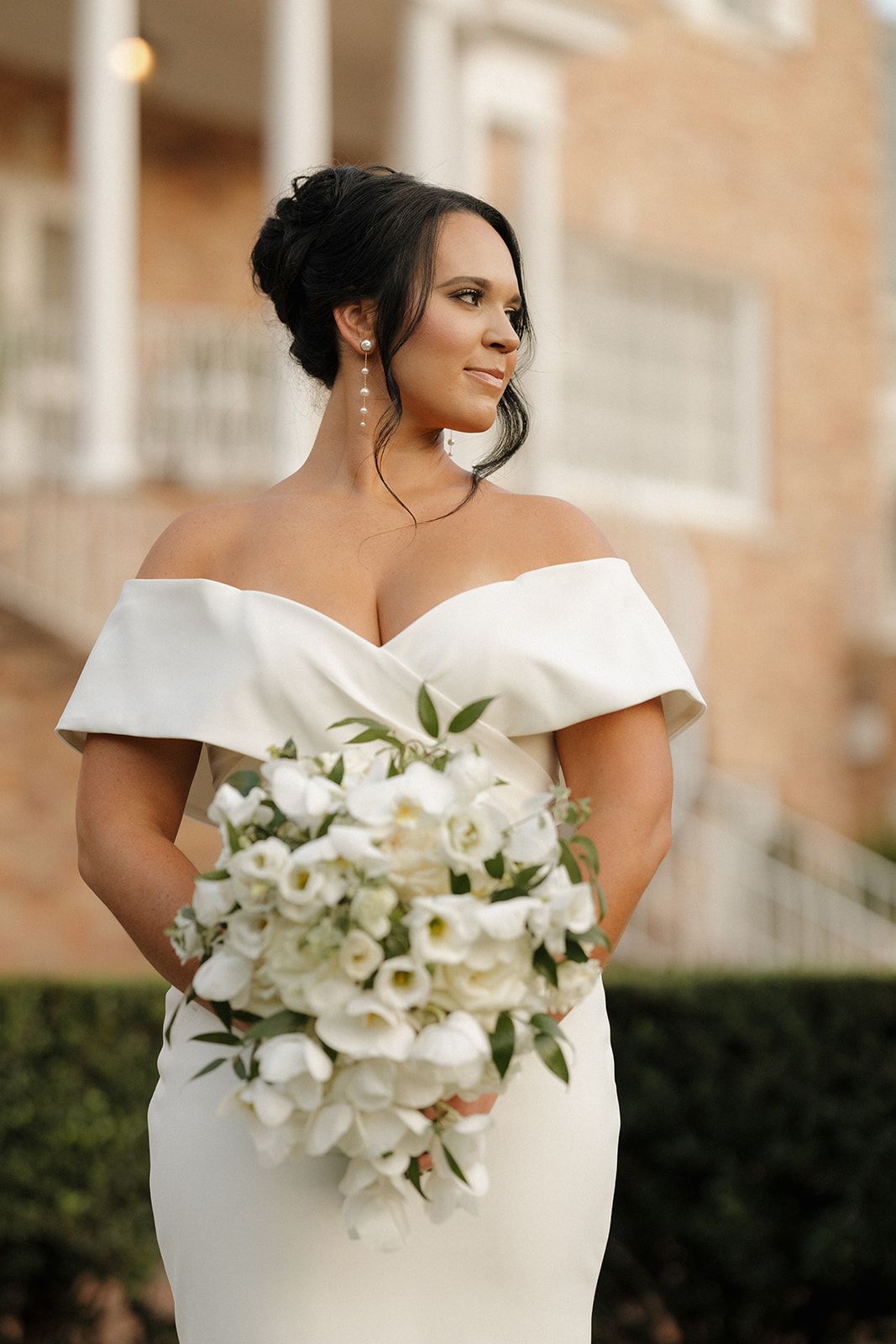 Bride in elegant off-shoulder white wedding dress holding beautiful white lily and orchid bouquet against brick building.