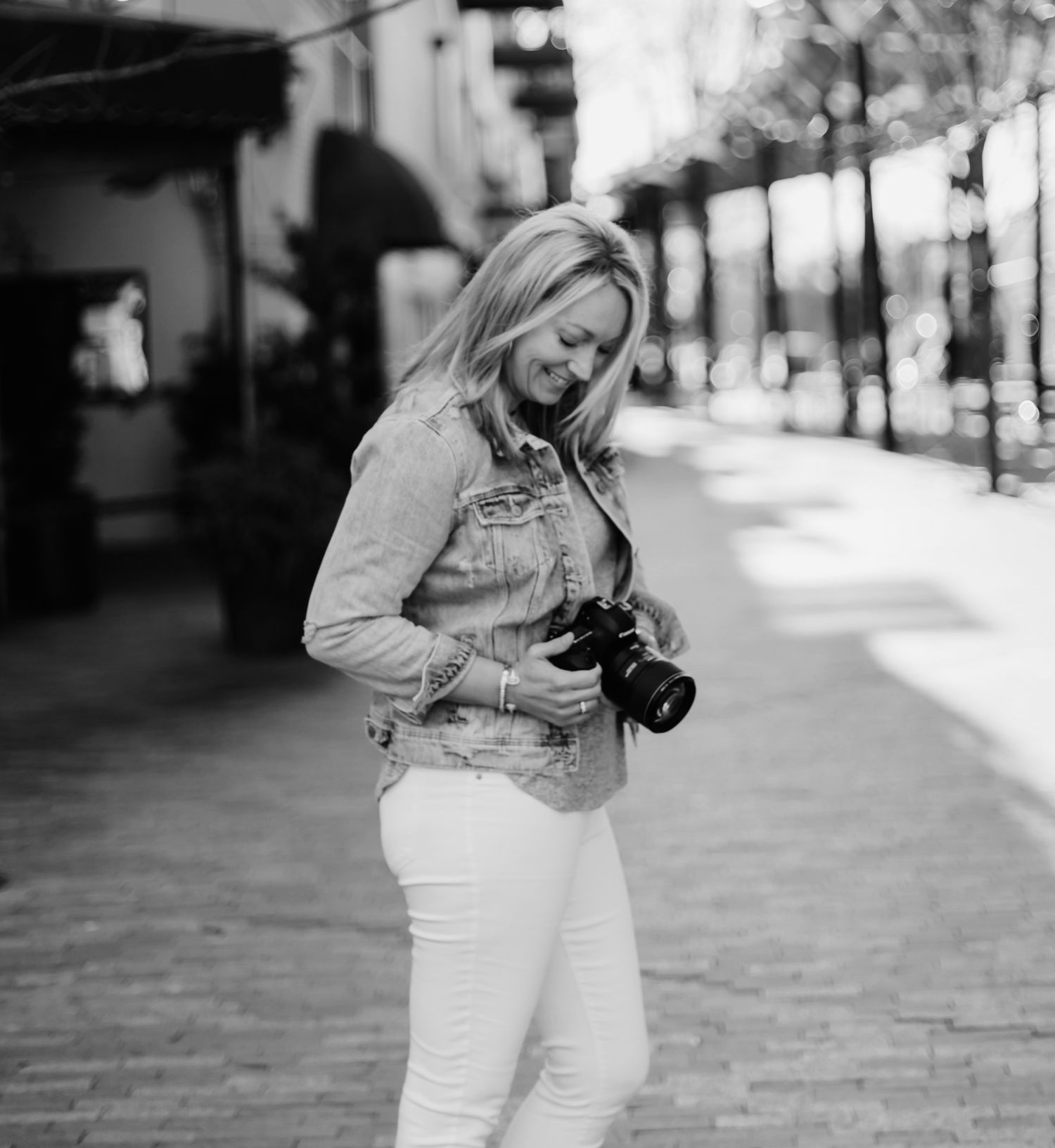 Person in white outfit examining digital camera while walking on urban sidewalk in black and white photograph.