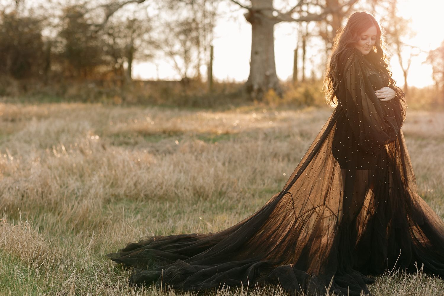 A silhouette stands in a golden field at sunset wearing a flowing black dress with dramatic long sleeves.