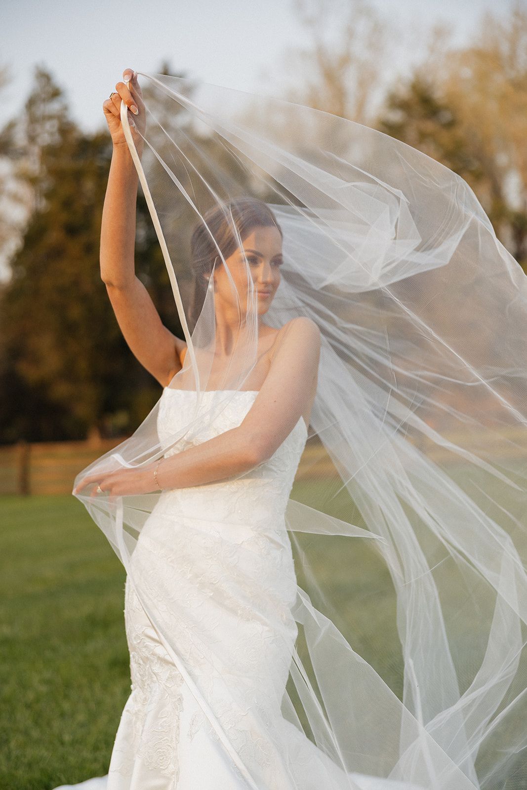 A bride in a white wedding dress stands in a field while her flowing veil billows dramatically in the breeze.