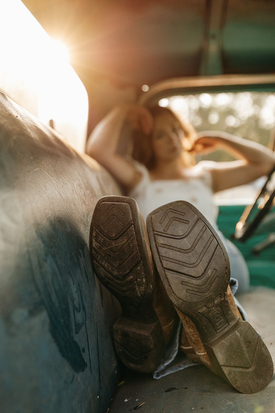 At sunset, muddy boots rest on a car dashboard as someone sits reclined in the driver's seat with warm sunlight streaming in.