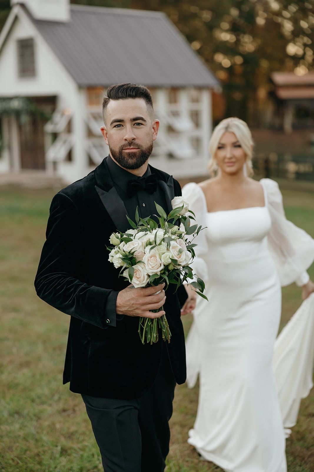 Wedding couple poses outdoors with white houses and autumn scenery in the background.